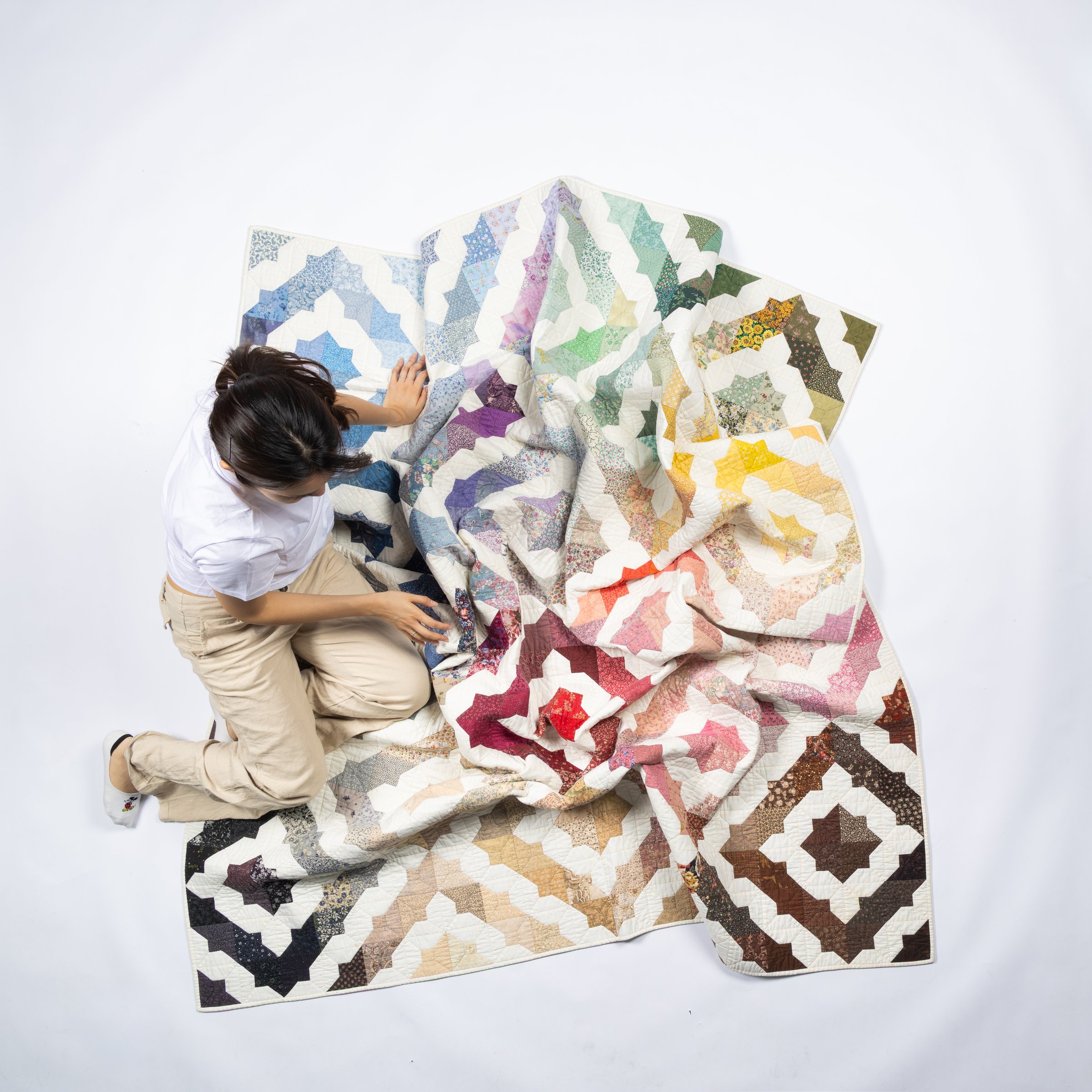 A person kneeling beside a large quilt, arranging the quilt by hand in a clean studio space.